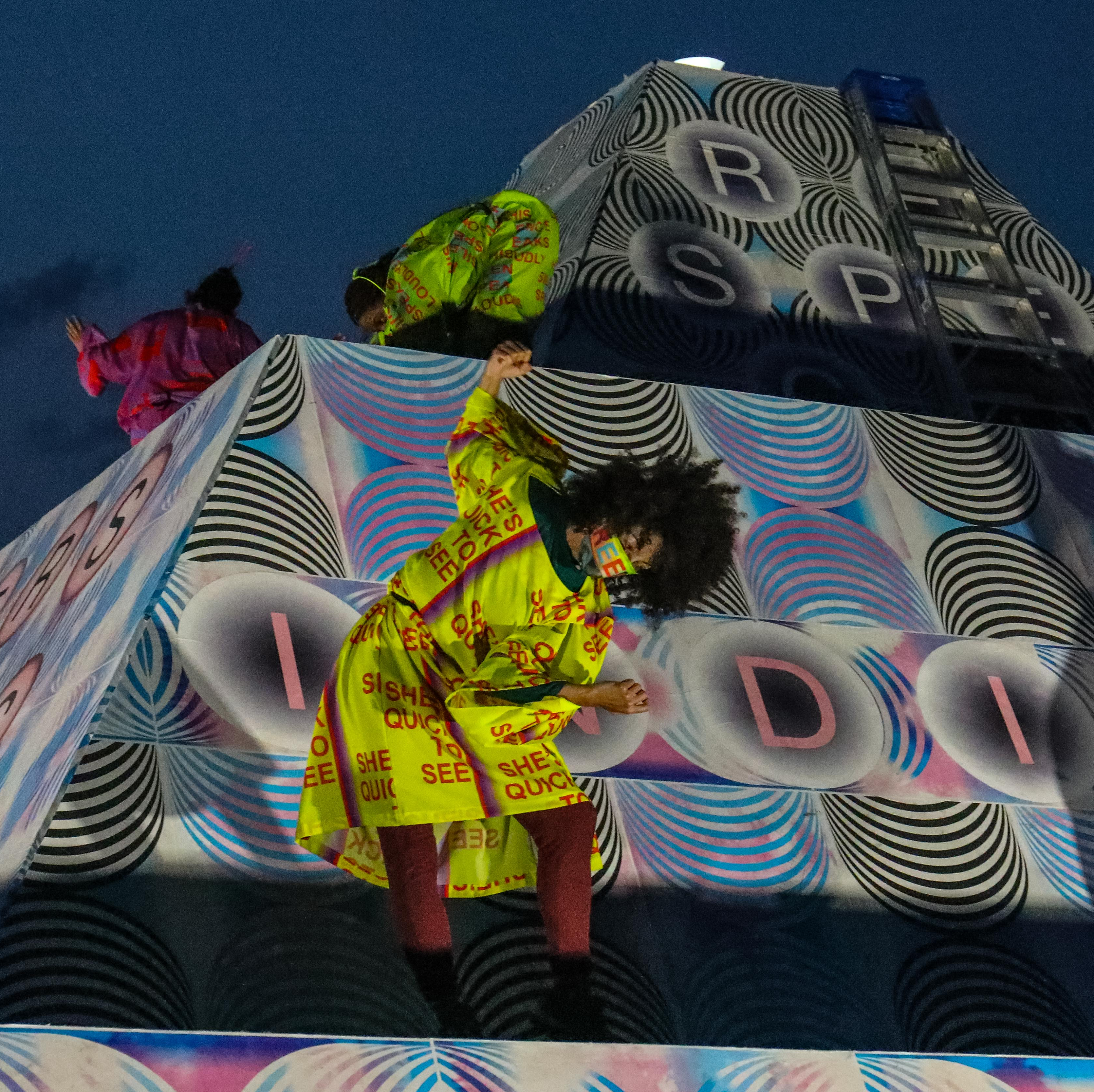 A woman in yellow dancing in motion on top of a sculpture.