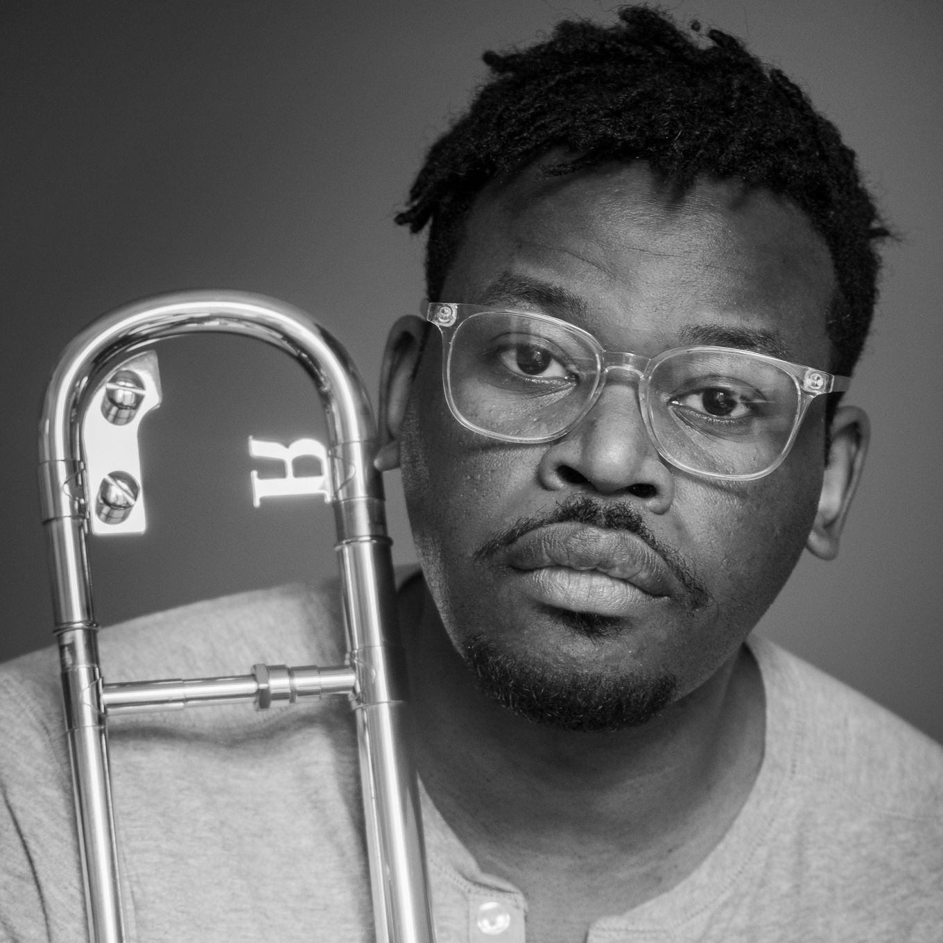 A black and white photo of a black man with glasses holding a trombone.