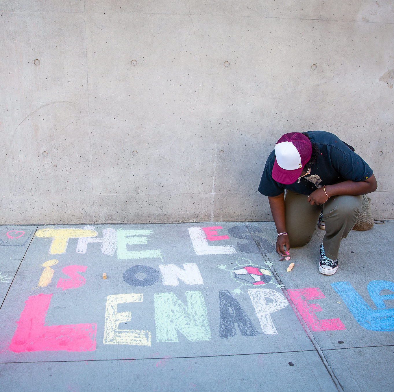 A sidewalk with a man kneeling and drawing in chalk "THE LES IS ON LENAPE LAND" 