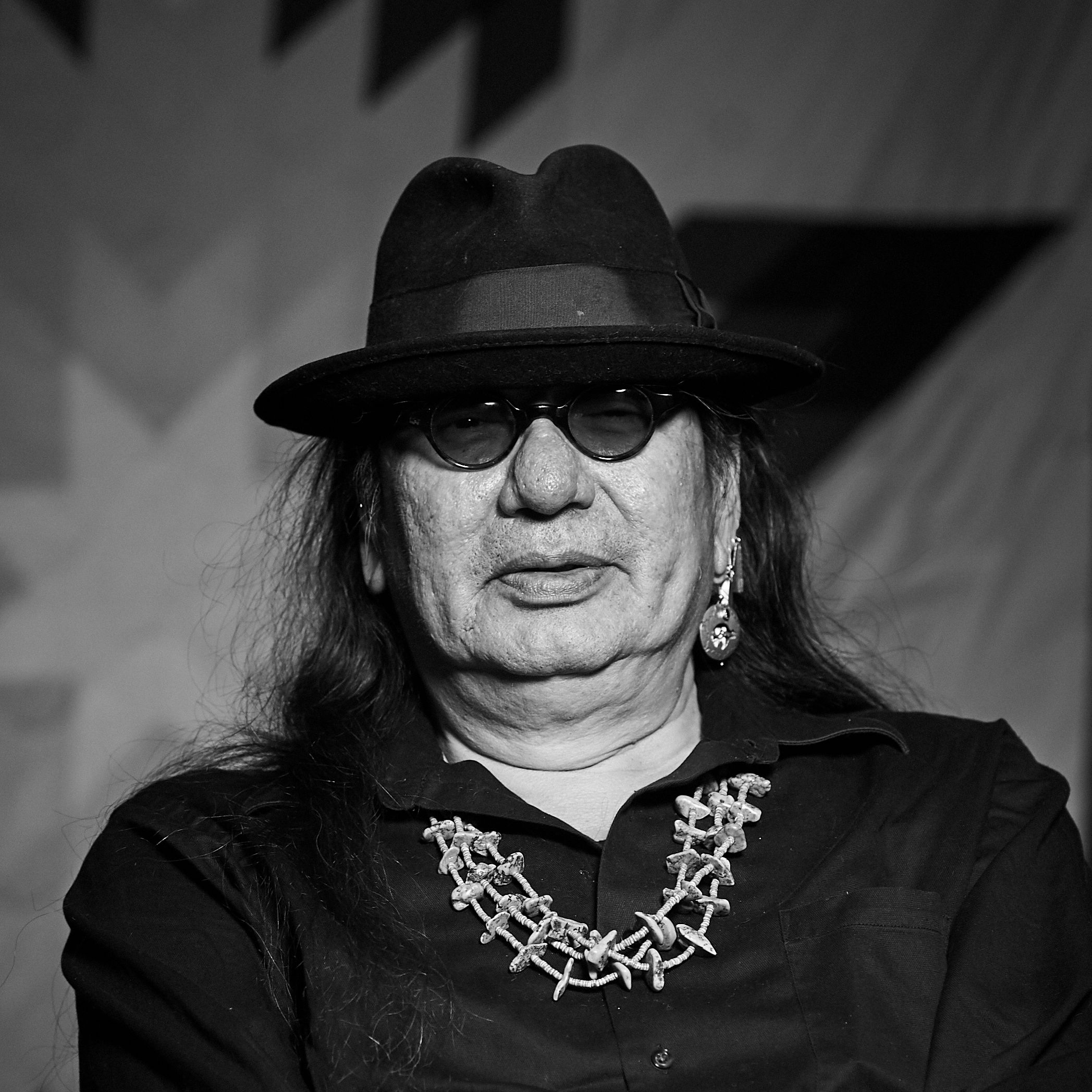 A black photo of a man with long hair, wearing glasses and a hat.