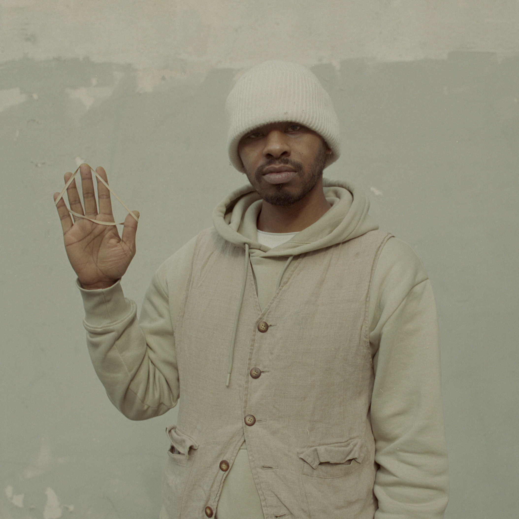 A black man in beige, wearing a white beanie with his right hand up.