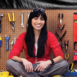 A woman with long hair and a red shirt, sitting cross-legged in front of a tool wall.