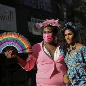 Two women, in a pink dress and a blue dress, holding a rainbow-colored fan. 